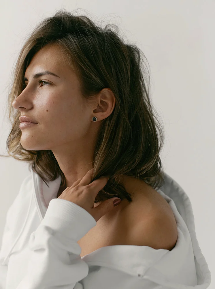 A close-up profile of a light-skinned woman with shoulder-length, slightly tousled dark brown hair. She has a neutral expression and is looking to her right. She wears a simple white loose-fitting top and a small dark stud earring on her left earlobe. Her smooth, clear skin and hand gently touching her neck and hair are highlighted. The background is a plain light gray/white, creating a soft, minimal backdrop that draws focus to the subject. - Chin Augmentation in New York and New Jersey