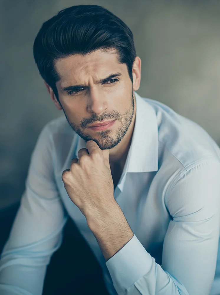 A close-up of a light-skinned man with short, dark hair. He wears a light-blue button-down dress shirt. His expression is thoughtful, with his head slightly tilted to the right and his right hand resting on his chin. His gaze is directed slightly to the right of the frame. The background is a muted greyish-taupe color. - Chin Implants for Men in New York and New Jersey