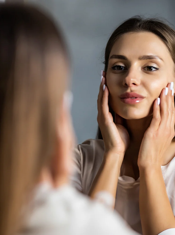 Close-up of a light-skinned woman with shoulder-length brown hair, looking at her reflection in a mirror. She is touching her face with both hands, visible from the wrists up, and her expression is pleasant and thoughtful. Soft, natural light illuminates her face. She is wearing a light cream-colored or beige blouse, and the background is a muted gray, slightly out of focus. Her reflection, including her hair and upper body, is visible in the mirror. - Dermabrasion in New York and New Jersey