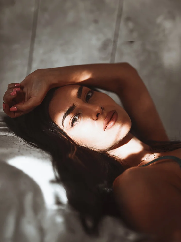 A woman with dark hair lies in bed, her head and upper body visible. Her arms are draped over her head, and her gaze is directed slightly forward. Soft lighting casts gentle shadows on her face and upper body, highlighting the texture of her skin. The background features a light gray textured surface, likely a wall or headboard. The white bed linens are mostly out of focus. A small gold or silver necklace and pinkish-nude nail polish on her fingernails are visible. - Facial Fat Grafting in New York and New Jersey