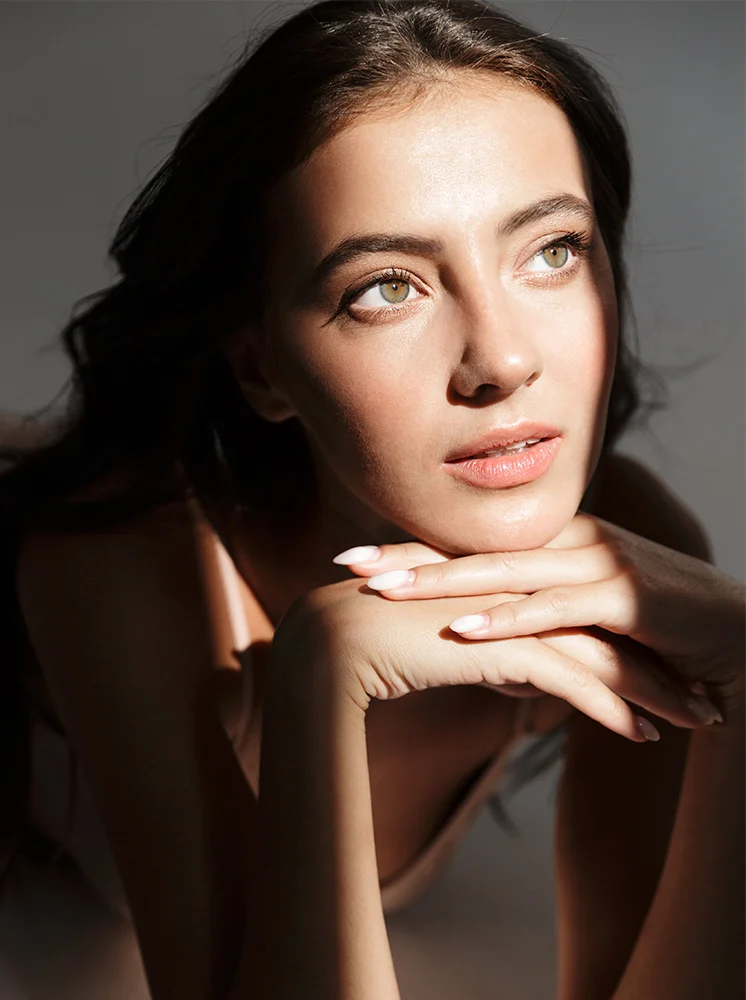 A woman with long, dark brown hair rests her head on her hands, gazing upward and slightly to her right with a contemplative expression. She has light, neutral-toned skin, light-olive/green eyes, and light pink lips. Her hands are stacked under her chin, with pale off-white nails visible. Strong shadows accentuate her facial features, with a gray-toned background adding depth. A soft pink/light-beige top is partially visible beneath her hands. - Gender Affirmation FAQs in New York and New Jersey