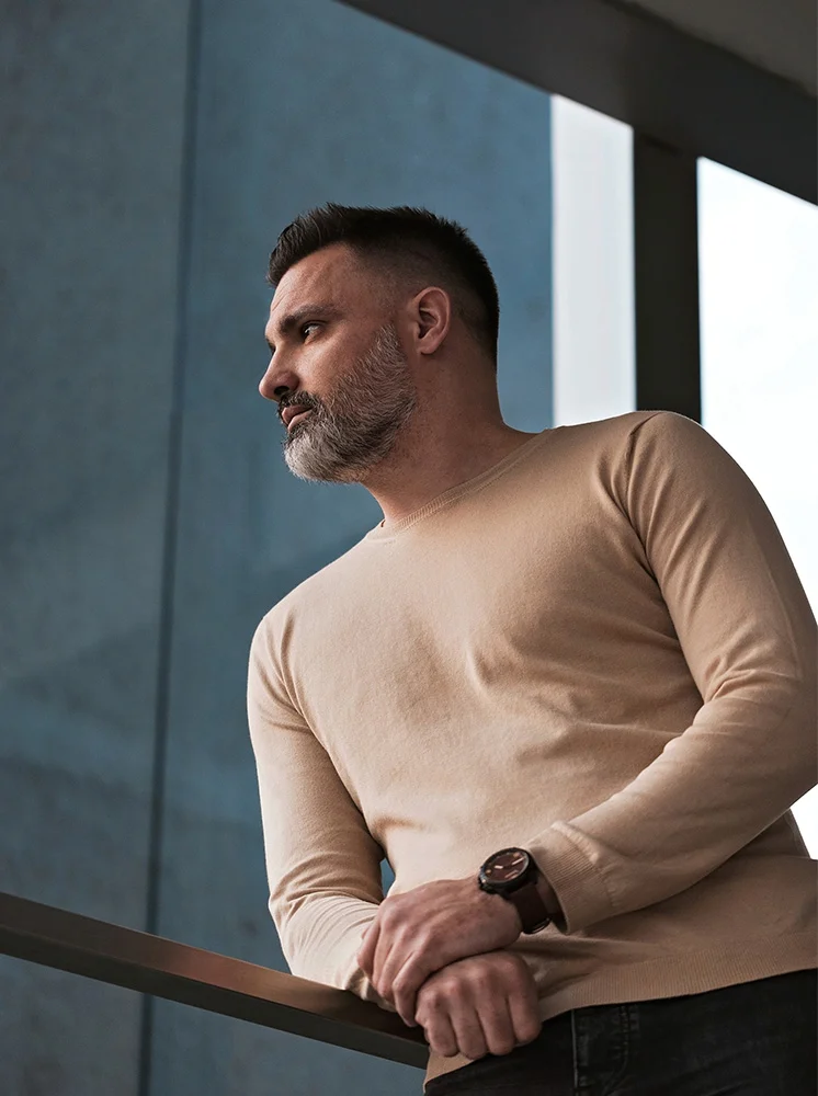 A man with short, dark hair and a gray beard is leaning against a metal railing, seen from the mid-chest up. He wears a light beige, long-sleeved top and has a contemplative expression, gazing upward and slightly to his right. His hands are clasped together, and a dark brown watch is visible on his left wrist. The background features a modern, muted blue-gray wall or building facade with soft shadows. A portion of the metal railing is visible, and the lighting is soft, enhancing the overall atmosphere. - Gynecomastia in New York and New Jersey