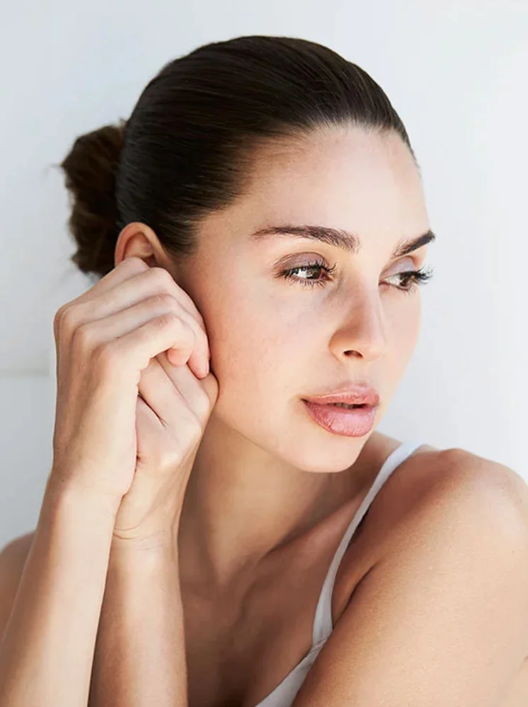 A close-up of a woman with shoulder-length dark brown hair styled in a low bun. She has a light complexion, defined eyebrows, and neutral makeup with rosy nude lips. She gazes slightly to her right with a thoughtful or serene expression. Her hands rest gently on her face and neck, with one hand near her ear. She wears a light cream-colored sleeveless top. The background is a plain, light gray-white wall, with soft lighting highlighting her features and creating a natural, soft aesthetic. - Morpheus8 in New York and New Jersey