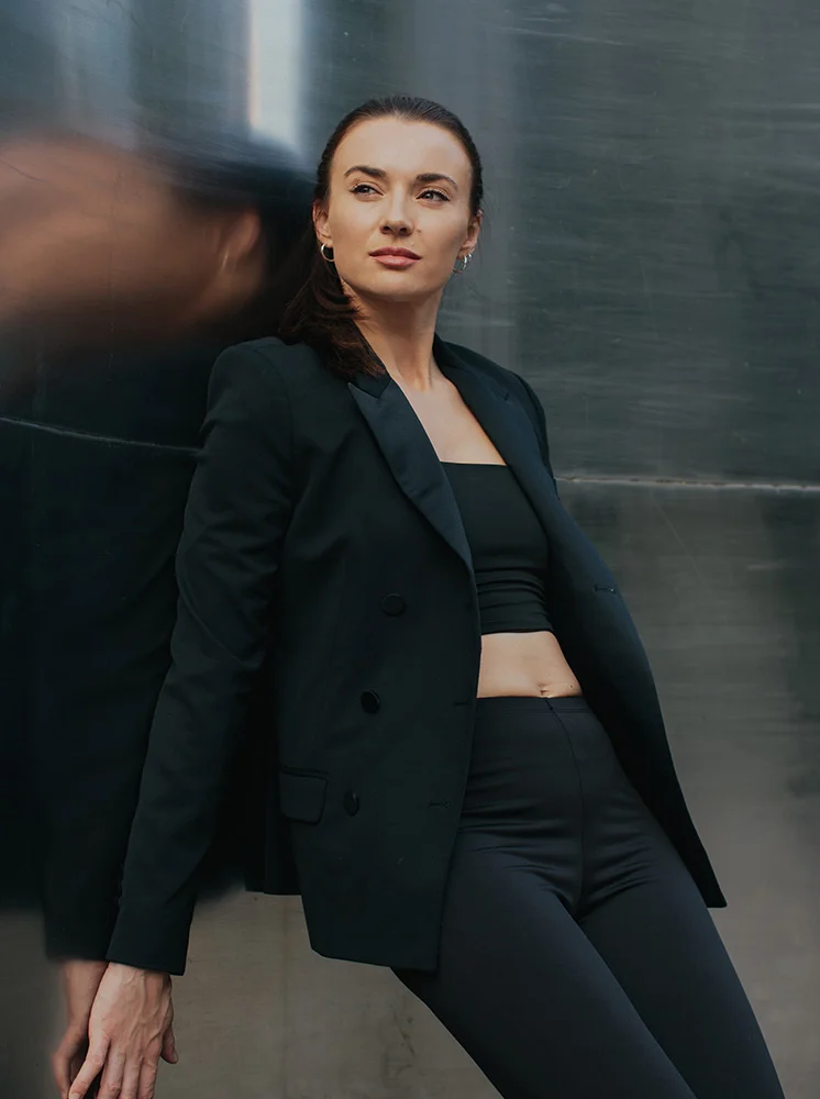 A woman leans against a gray metal surface, wearing a black blazer over a black cropped top and pants. The blazer features double-breasted buttons. Her shoulder-length brown hair is pulled back in a ponytail. With a thoughtful expression, she gazes off to her right. A blurred reflection of the woman and the wall appears behind her. Natural lighting gives the image a sophisticated tone. - Nipple Reconstruction in New York and New Jersey