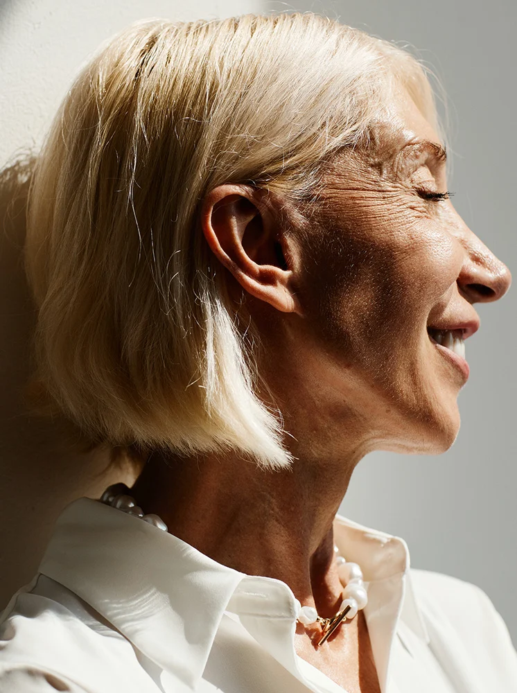 Close-up profile of a woman with short, light blonde hair, seen from the mid-chest up. She has visible wrinkles and fine lines, with a relaxed, serene expression and a slight smile. She wears an off-white, collared top—possibly silk or crepe—and a choker-style necklace with off-white pearls and gold-toned accents. - Surgical Approaches to lymphedemia in New York and New Jersey
